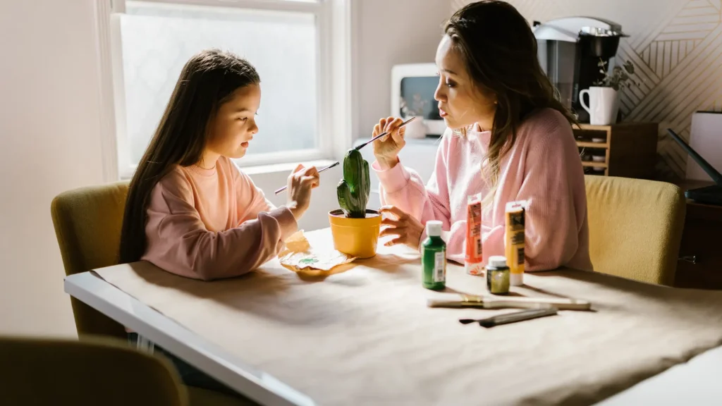 Mother and daughter painting a small potted plant together at a table.
